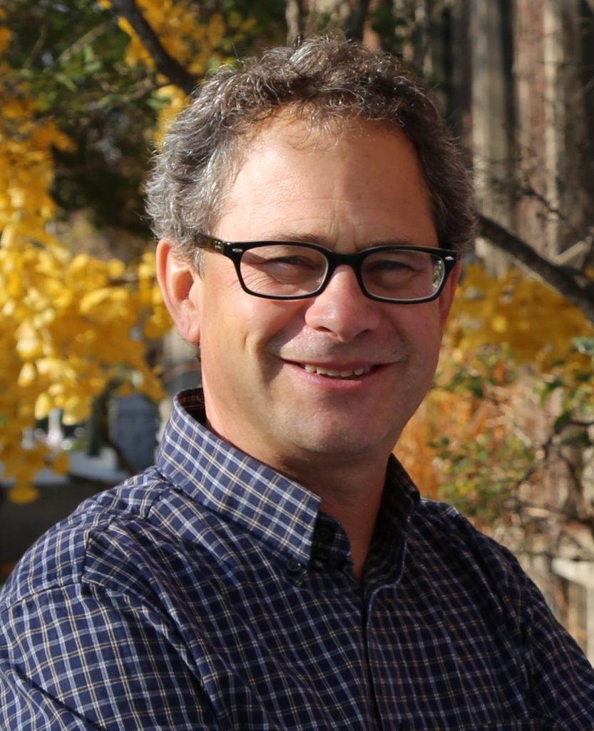 head shot of Jeff Brook, blue checked button down shirt; foliage in background