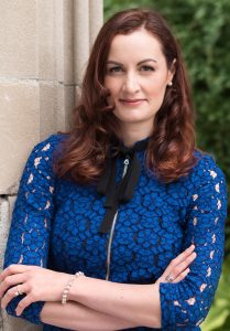 Laura Rosella in blue top with arms folded leaning against cement wall outdoors