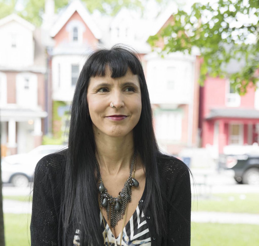 Prof. Suzanne Stewart in front of downtown Toronto houses wearing black jacket and necklace
