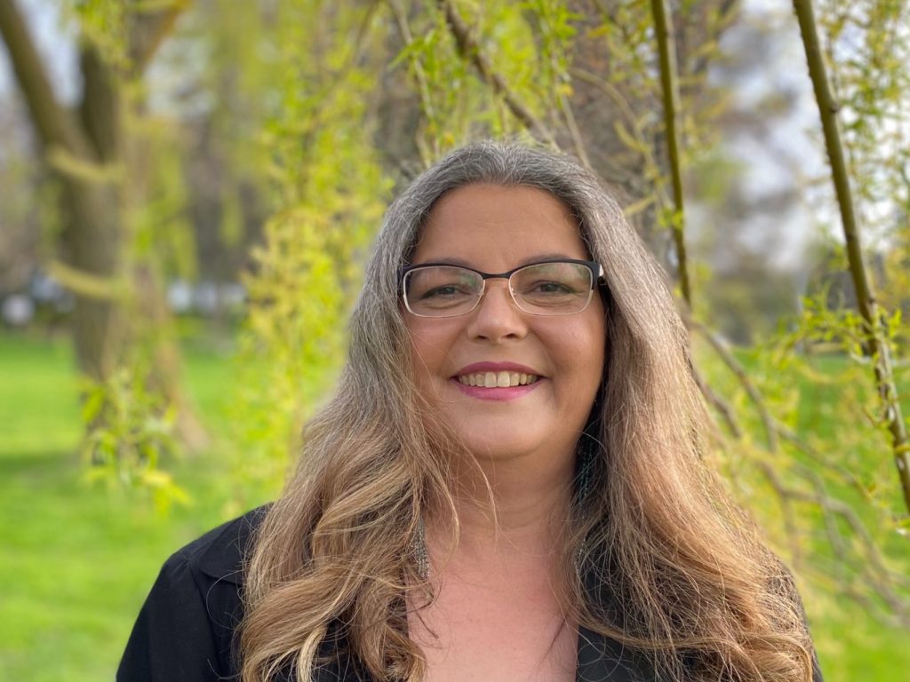head shot of Assoc. Prof. Angela Mashford-Pringle with long hair, glasses, open-necked top, spring leaves in wooded background