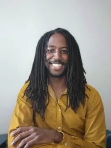 Cornel Grey, smiling, arms folded, mustard coloured long sleeved shirt