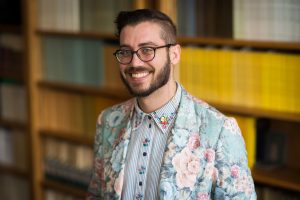 C Dalrymple-Fraser standing in front of a bookcase, smiling. And a slightly fuller image description has been: C Dalrymple-Fraser, a white person with a short hairstyle and facial hair, is standing in a brightly lit office in front of a bookcase. They have round glasses and are wearing a floral-patterned blazer. They are facing the camera and smiling.