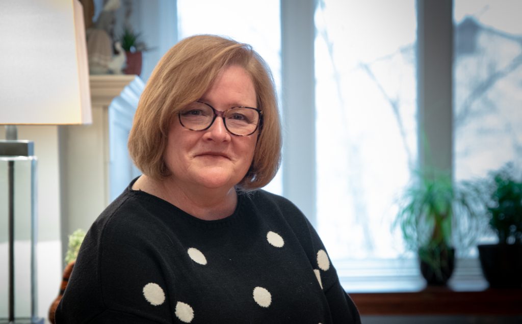 Prof. Carol Strike in black sweater with white polka dots, glasses, chin length bob, in front of window in her home