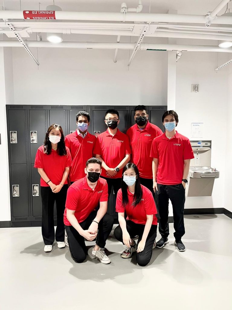 five students standing and two crouching in front, all in matching red polo shirts and black pants, all wearing masks