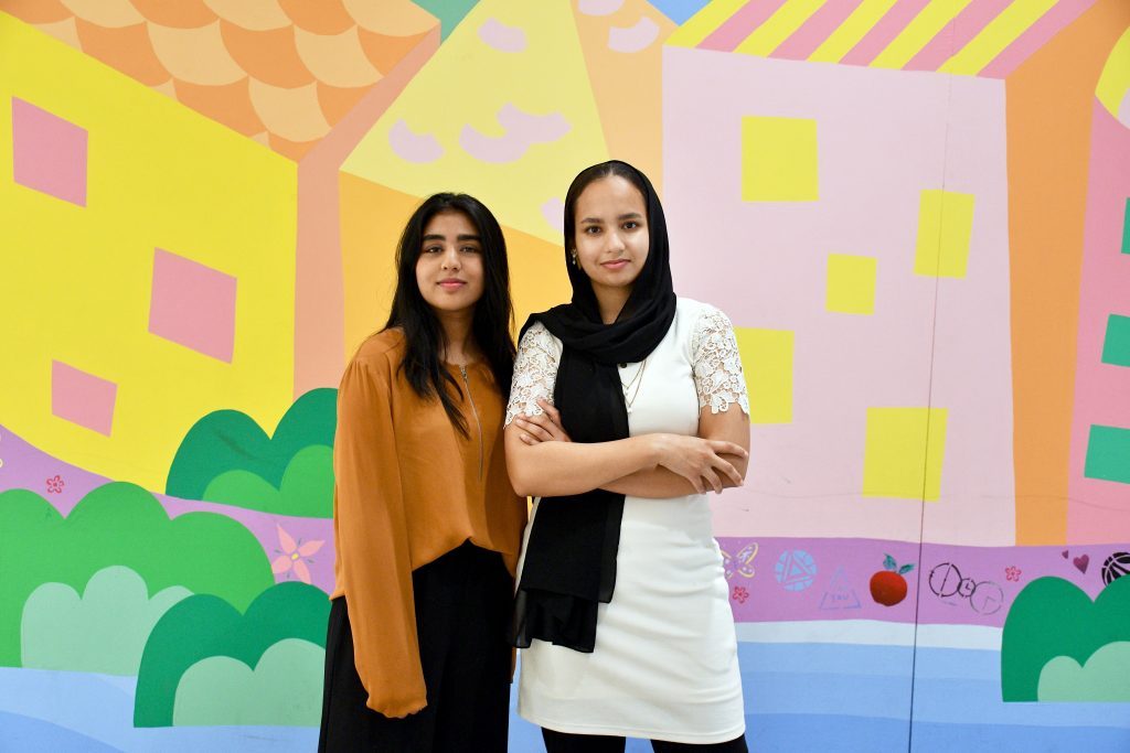 The two undergrad students pose in front of a mural made by members of The Neighbourhood Organization.