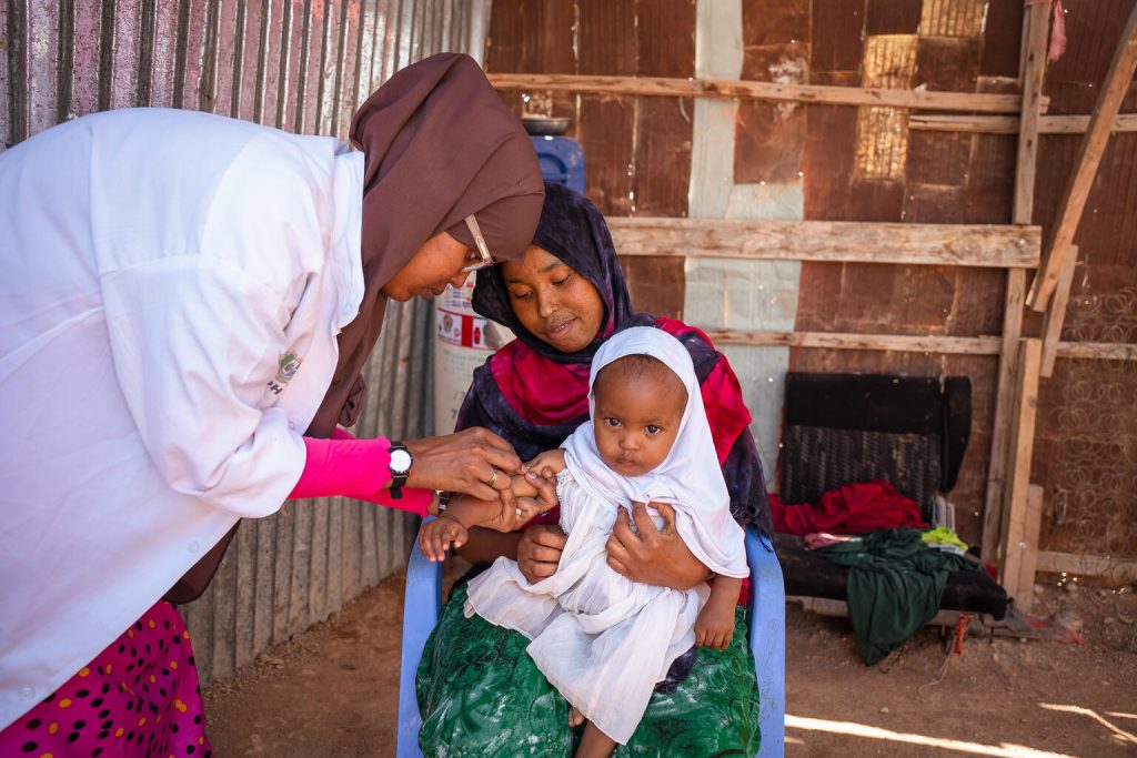 In in Puntland, Somalia, 24-year-old clinical health worker and social worker Kowther Abdikadir vaccinates Muniish Cadan Ismail, who sits on the lap of her mother, Zeynab Mohamed Farah, during a community outreach effort conducted as part of Jilab Health Centre’s ‘fixed temporary outreach’ immunization and health programme. (Grace Ekpu VII Photo, UNICEF)