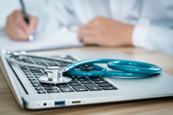 Stock photo of stethoscope on top of laptop keyboard. A doctor's hands write notes in the background.