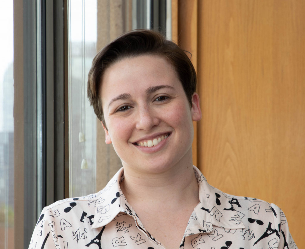 Headshot of Gilla Shapiro standing in front of a window and smiling