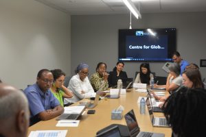 A group of people sitting around a large table in a board room. A screen is mounted on the wall.